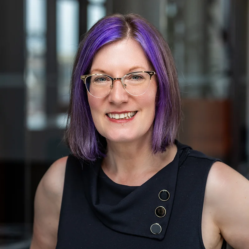 Headshot of Rebecca Colwell, smiling, with purple hair and gold-rimmed glasses.