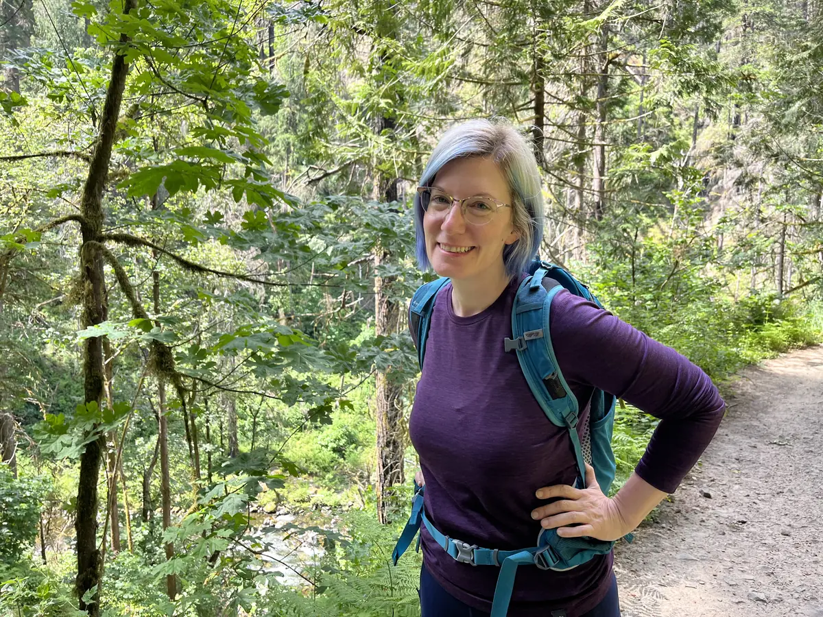 Rebecca smiling on a forested trail in hiking gear, with a creek below.